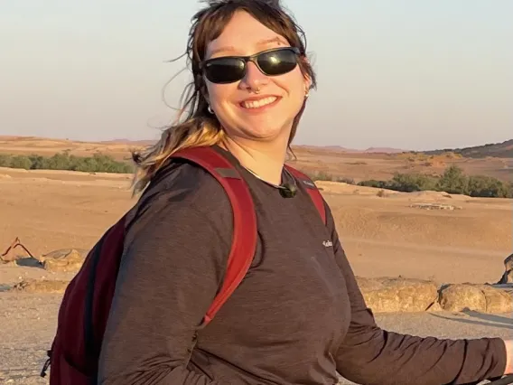 Avery stands on a path wearing a backpack and sweater. The background is sandy with sparse vegetation.