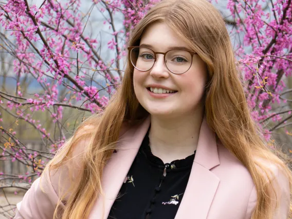 Katelyn stands in front of a tree with dark pink blooms. She wears a light pink blazer and smiles broadly.