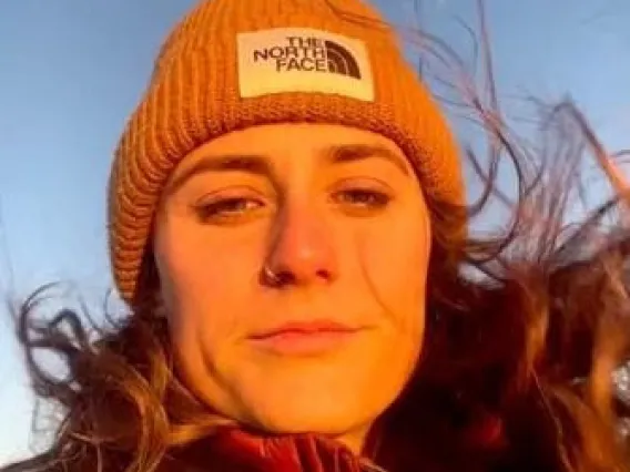 Caitlin looks down in this close-up photo. The wind blows her hair around and she wears a warm hat. The setting sun bathes her face in golden light. 