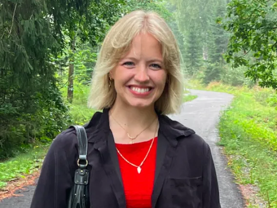 Dakota stands in the middle of a rural road that is surrounded by lush vegetation. She wears a jacket and a red top. 