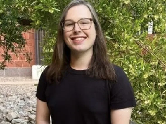 Griffin sits on a bench, smiling with her hands in her lap. Foliage and a brick building fill the background.