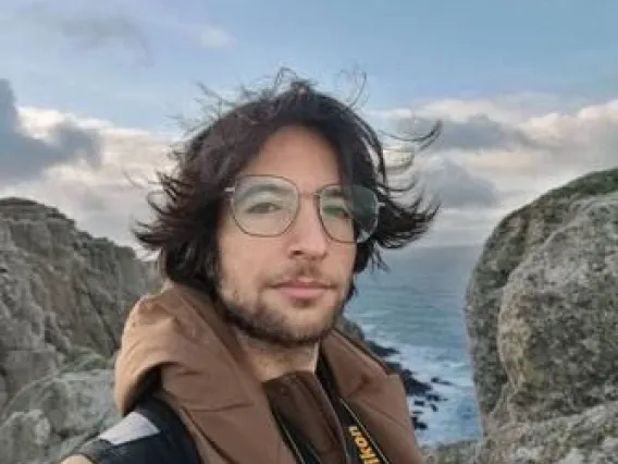 Michael stands on a hill with the wind blowing his hair. A rocky beach and a cloudy sky fill the background. 