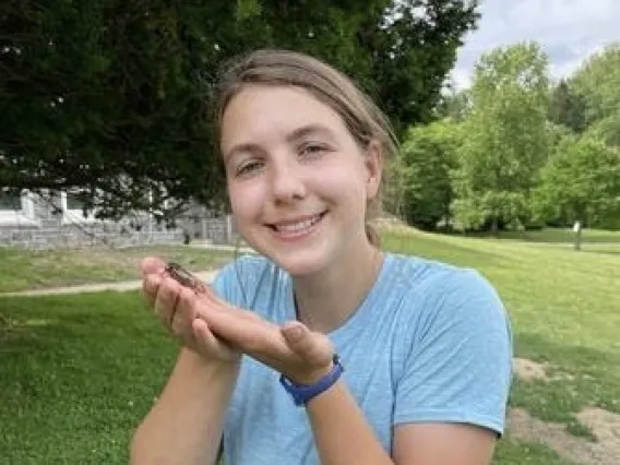 Poppy stands on a grassy hill and cheerfully presents a large insect. Trees fill the background.