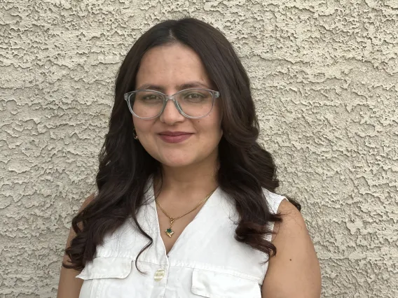 Sallu stands against a stucco wall. She wears a white sleeveless top.