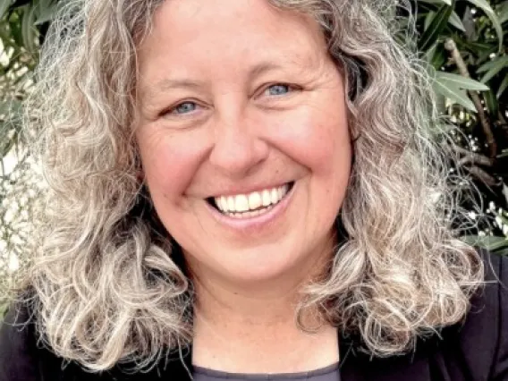 Betsy stands in front of a tree. She wears a black jacket and grey top. 