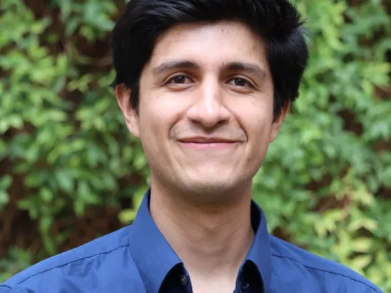 Christian stands outside with foliage filling the background. He wears a blue button up top. 