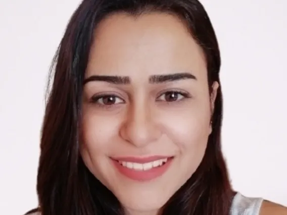 Ghiwa stands in front of a plain, light background. She wears a white sleeveless top and smiles warmly. 