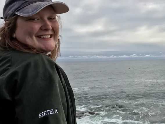 Jes smiles widely as she poses for a photo. She stands on a boat with choppy water in the background. 