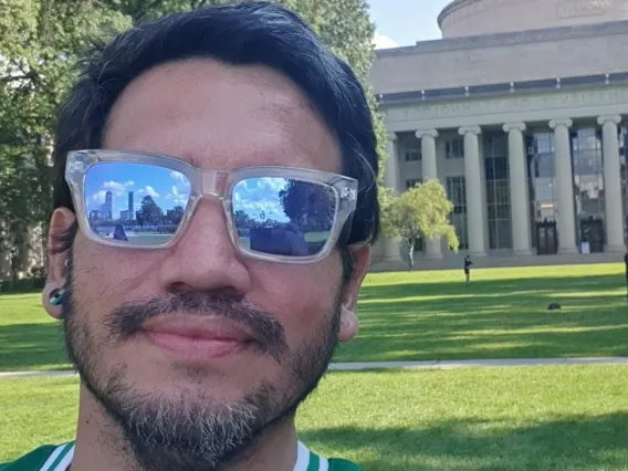 Pablo poses in front of a Neo-classical building and lawn. He wears sunglasses and a green shirt.