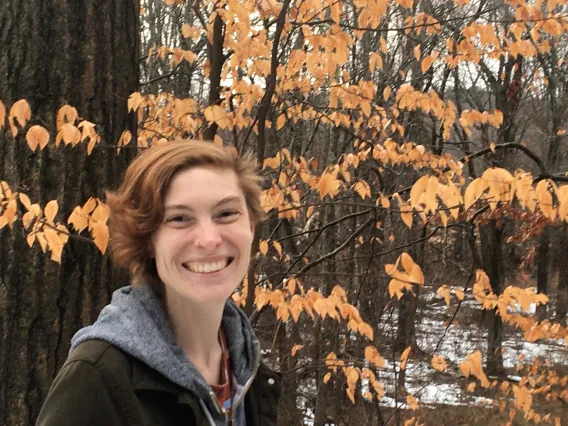 Rebecca stands in a forest. The leaves are orange and there are patches of snow on the ground. She wears a jacket and sweatshirt to stay warm. 