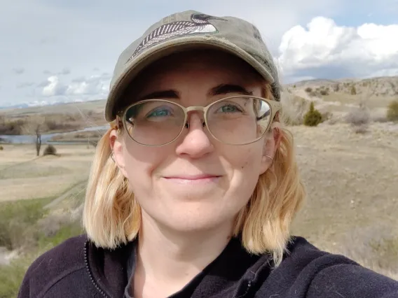 Savannah poses for a photo outside. They wear a cap and jacket.  A stream and dry grassland fill the background. 