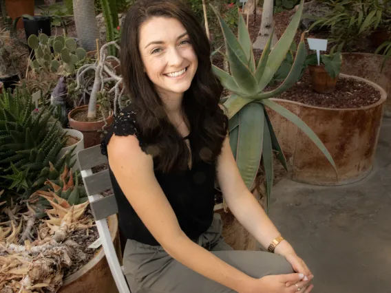 Taylor sits on a folding chair with her hands in her lap. Desert plants in pots fill the background. 