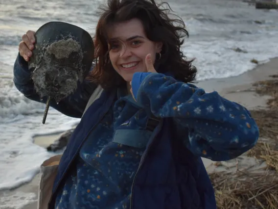 Meredith stands on a windy beach holding a stingray. She gives a big smile and thumbs up for the camera.