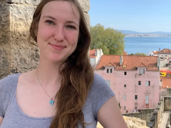 Zoe stands in front of a cut rock opening. Ocean and colorful buildings fill the background. 