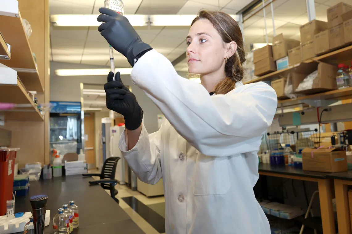 Caitlin works in the lab, looking at a test tube while wearing gloves and lab coat