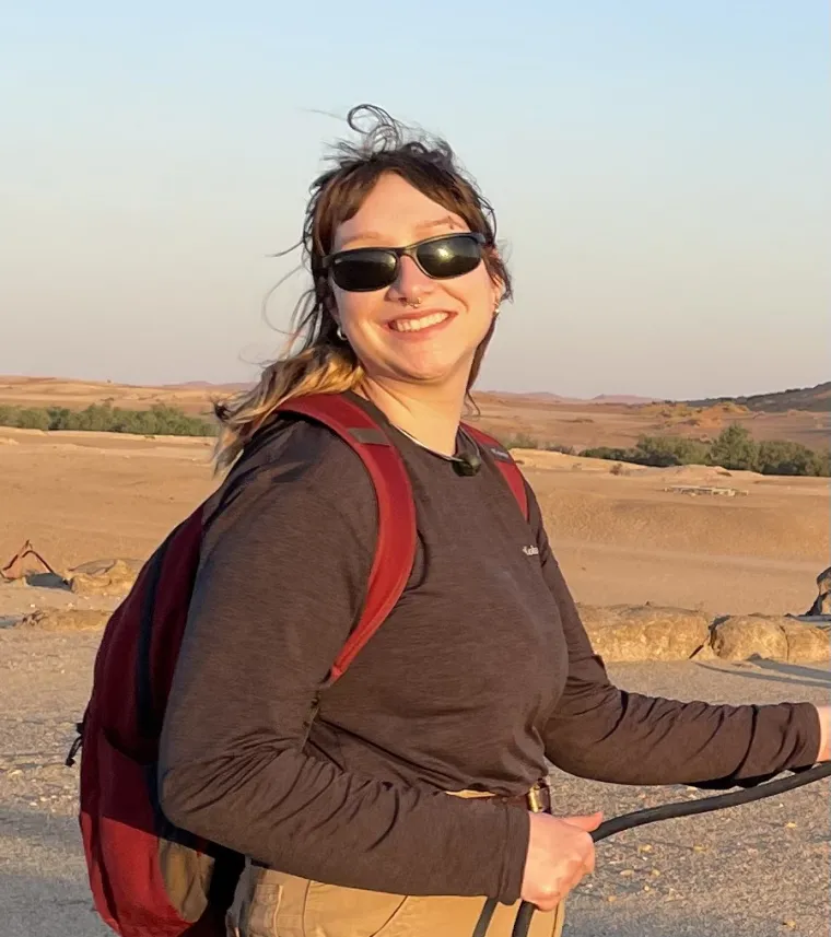 Avery stands on a path wearing a backpack and sweater. The background is sandy with sparse vegetation.