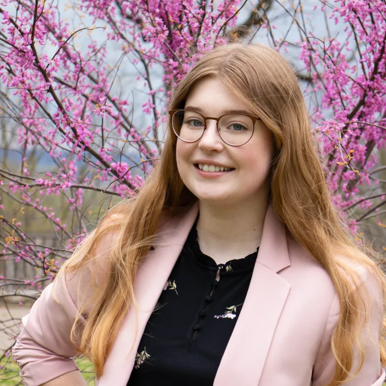 Katelyn stands in front of a tree with dark pink blooms. She wears a light pink blazer and smiles broadly.