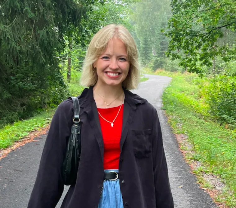 Dakota stands in the middle of a rural road that is surrounded by lush vegetation. She wears a jacket and a red top. 