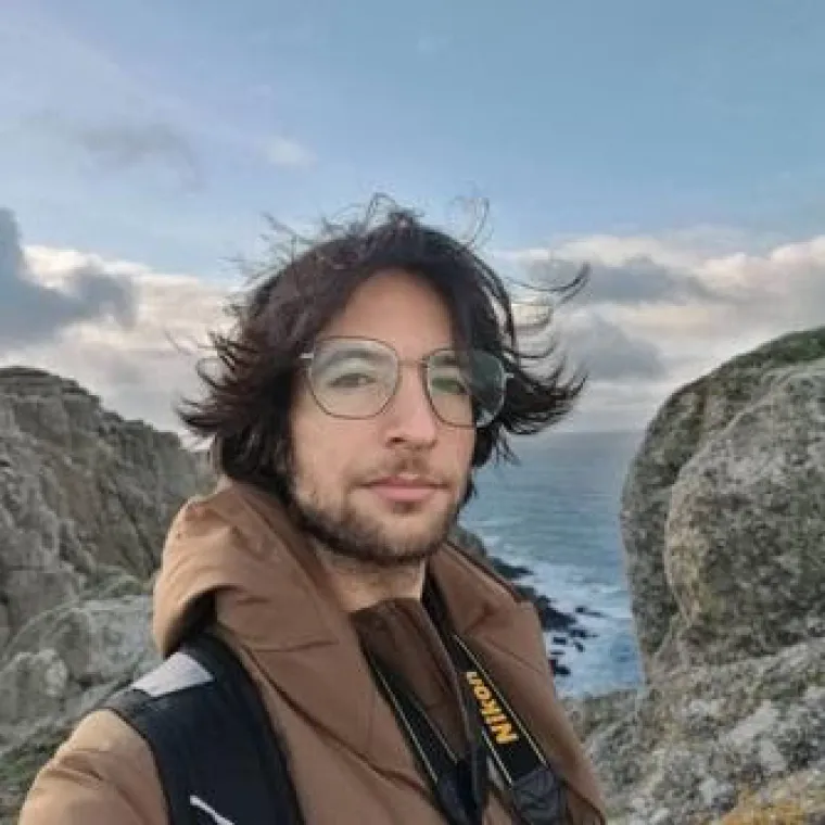 Michael stands on a hill with the wind blowing his hair. A rocky beach and a cloudy sky fill the background. 