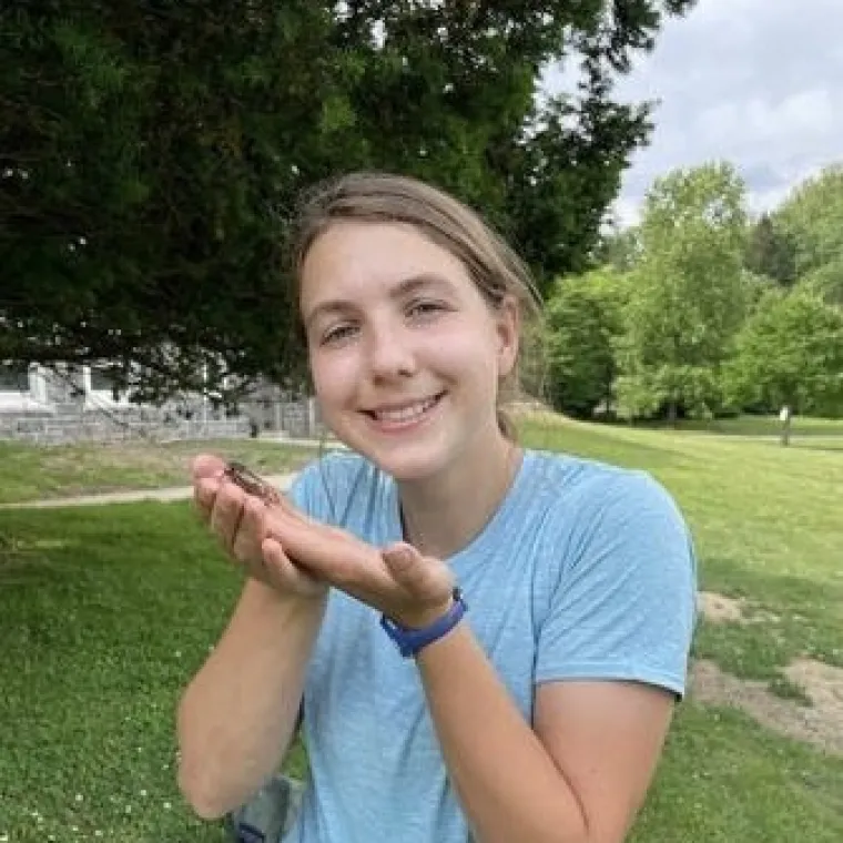 Poppy stands on a grassy hill and cheerfully presents a large insect. Trees fill the background.