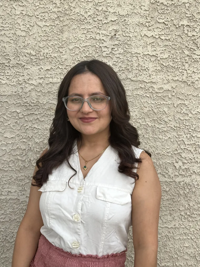 Sallu stands against a stucco wall. She wears a white sleeveless top.