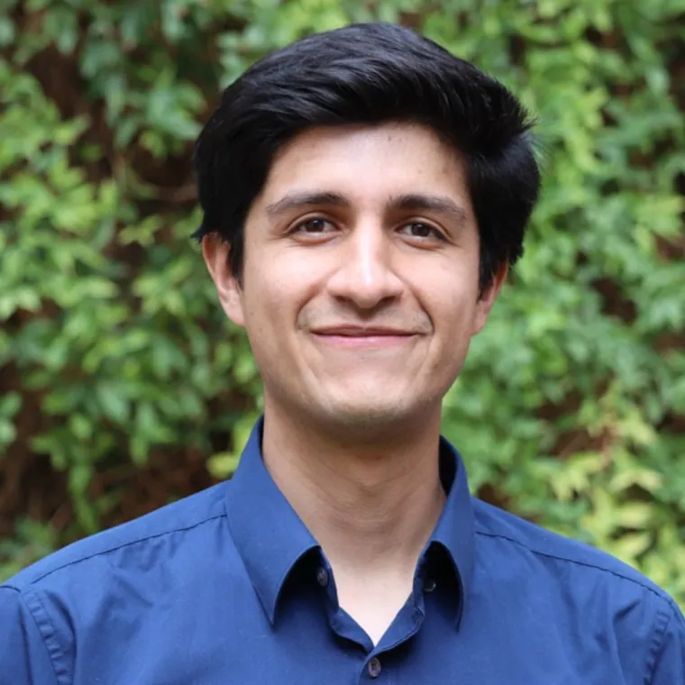 Christian stands outside with foliage filling the background. He wears a blue button up top. 