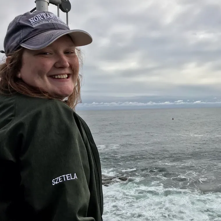 Jes smiles widely as she poses for a photo. She stands on a boat with choppy water in the background.