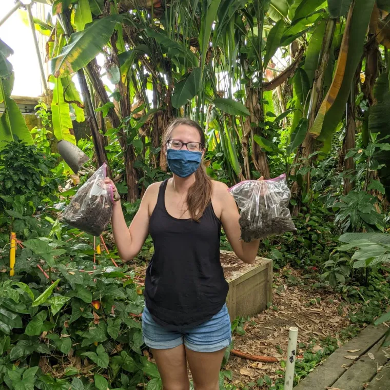 Meara stands holding soil samples in a tropical forest. She is masked and wears casual clothing. 