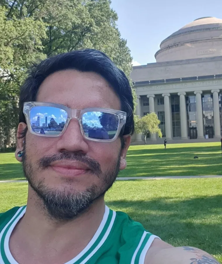 Pablo poses in front of a Neo-classical building and lawn. He wears sunglasses and a green shirt.