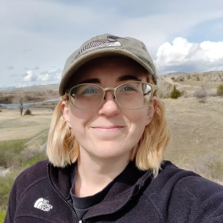 Savannah poses for a photo outside. They wear a cap and jacket.  A stream and dry grassland fill the background. 