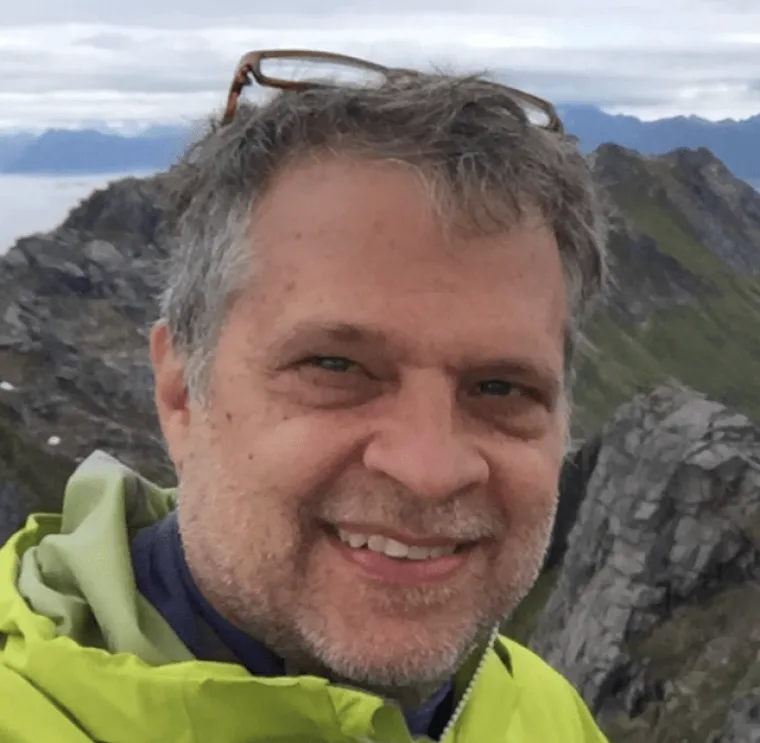 Scott takes a photo during fieldwork. He wears a yellow rain jacket and stands on a hill.