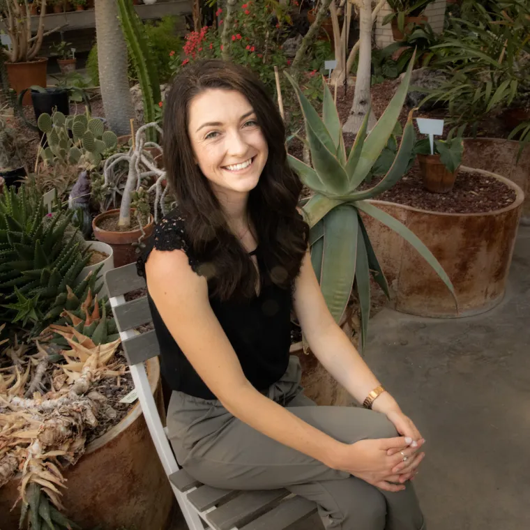 Taylor sits on a folding chair with her hands in her lap. Desert plants in pots fill the background.