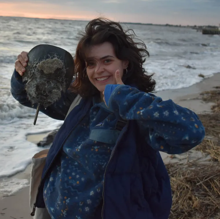 Meredith stands on a windy beach holding a stingray. She gives a big smile and thumbs up for the camera.