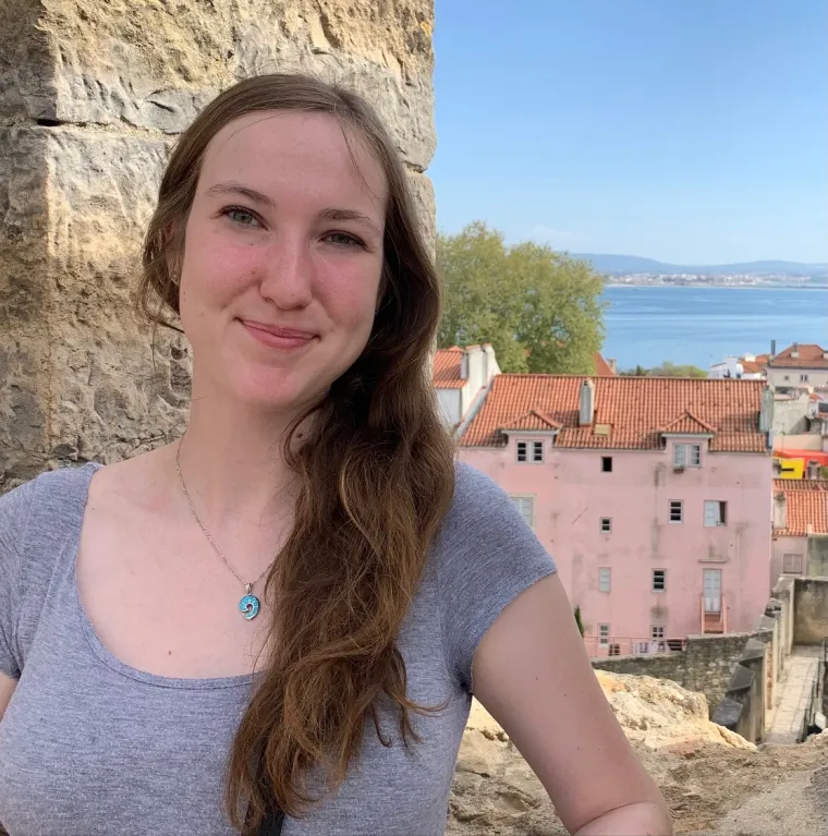 Zoe stands in front of a cut rock opening. Ocean and colorful buildings fill the background. 